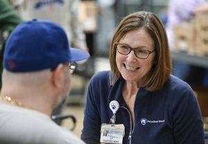 A nurse smiles at a man who is seen from behind wearing a baseball cap and glasses. The nurse has a jacket with a Penn State Health logo on it and is wearing a name badge and glasses.