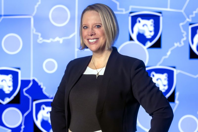 Dr. Jennifer Kraschnewski poses for a photograph, wearing professional attire. The background is a map with several Penn State Nittany Lion shield logos.