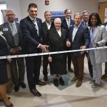 Ten people wearing professional attire pose for a photo in a hospital lobby area as they prepare to cut a ribbon with the “Penn State Health” logo.
