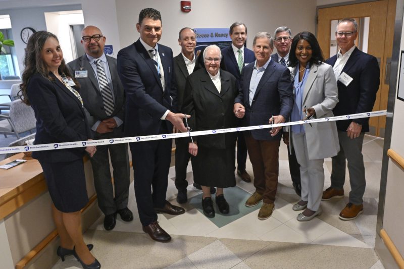 Ten people wearing professional attire pose for a photo in a hospital lobby area as they prepare to cut a ribbon with the “Penn State Health” logo.