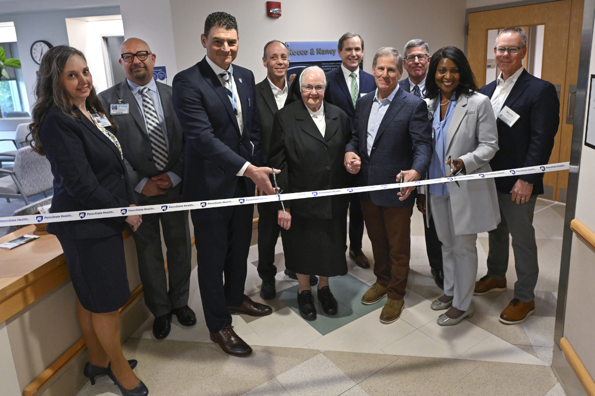 Ten people wearing professional attire pose for a photo in a hospital lobby area as they prepare to cut a ribbon with the “Penn State Health” logo.