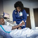 A nurse in a hospital room adjusts an IV line on a male patient lying in a bed, while monitoring equipment and a computer are visible in the background.
