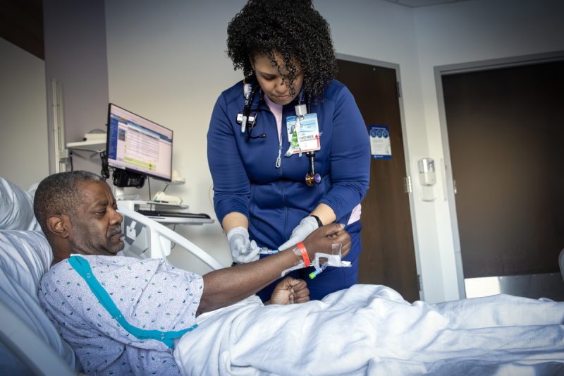 A nurse in a hospital room adjusts an IV line on a male patient lying in a bed, while monitoring equipment and a computer are visible in the background.