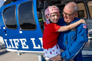 A man in a blue flight jumpsuit holds a young girl who’s wearing a helmet and smiling. They are directly in front of a Life Lion helicopter on which the side door is open.