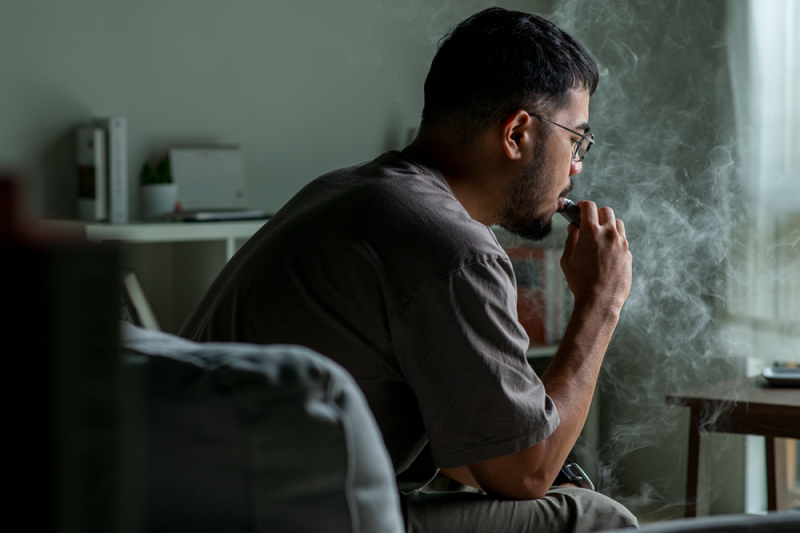 An individual sitting on a couch uses an e-cigarette.