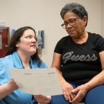 A doctor holds a piece of paper toward a woman patient who is seated on an exam table. The doctor is wearing a blouse with a lanyard around her neck. The woman patient is wearing glasses and a T-shirt with the “Guess” logo on it.