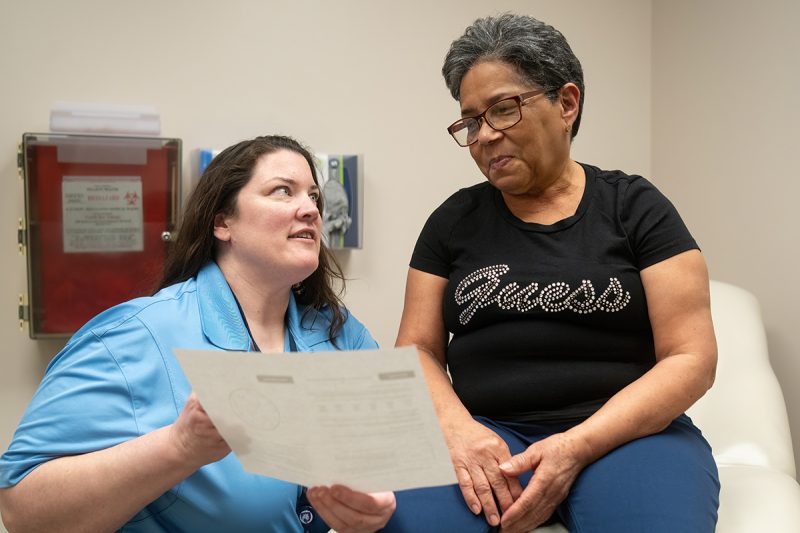 A doctor holds a piece of paper toward a woman patient who is seated on an exam table. The doctor is wearing a blouse with a lanyard around her neck. The woman patient is wearing glasses and a T-shirt with the “Guess” logo on it.