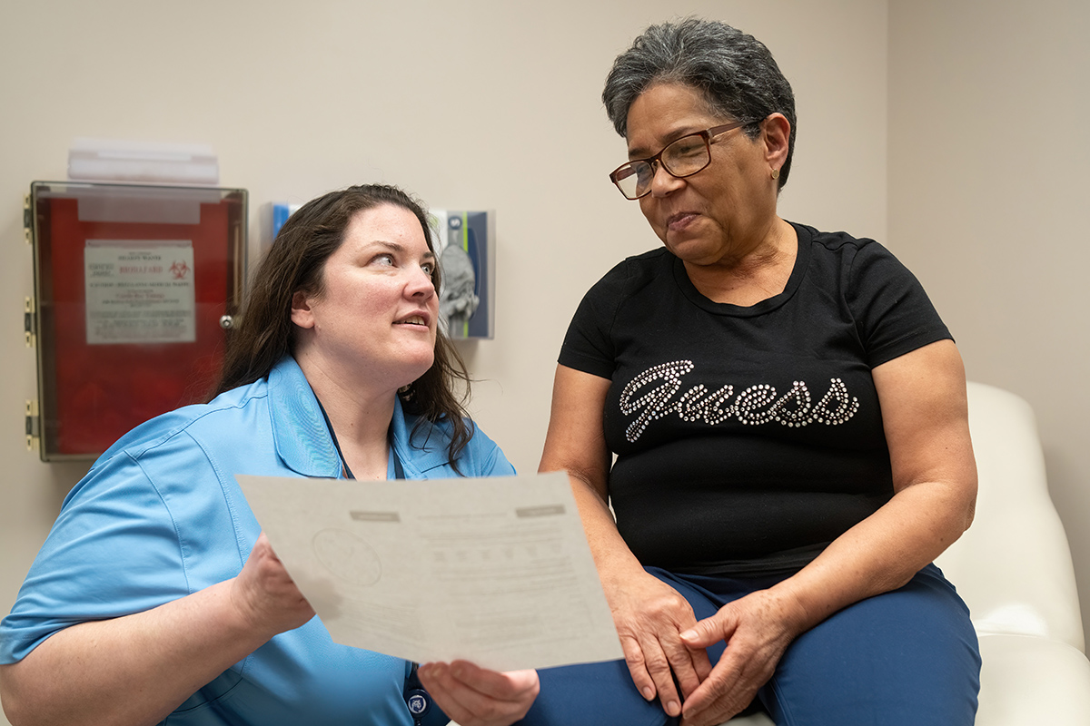 A doctor holds a piece of paper toward a woman patient who is seated on an exam table. The doctor is wearing a blouse with a lanyard around her neck. The woman patient is wearing glasses and a T-shirt with the “Guess” logo on it.