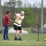 A man swings a golf club while wearing a gait belt and leg brace on his left leg. A man on the left holds his gait belt to give him balance support. A cane is to the right of the golfer.