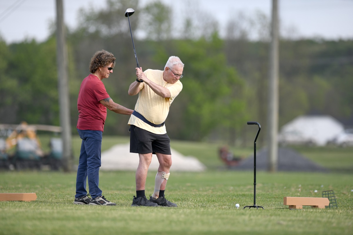 A man swings a golf club while wearing a gait belt and leg brace on his left leg. A man on the left holds his gait belt to give him balance support. A cane is to the right of the golfer.