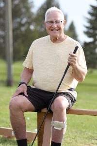 A man smiles while sitting on a bench at a golf course. He is holding a golf club and wearing a leg brace on his left leg.