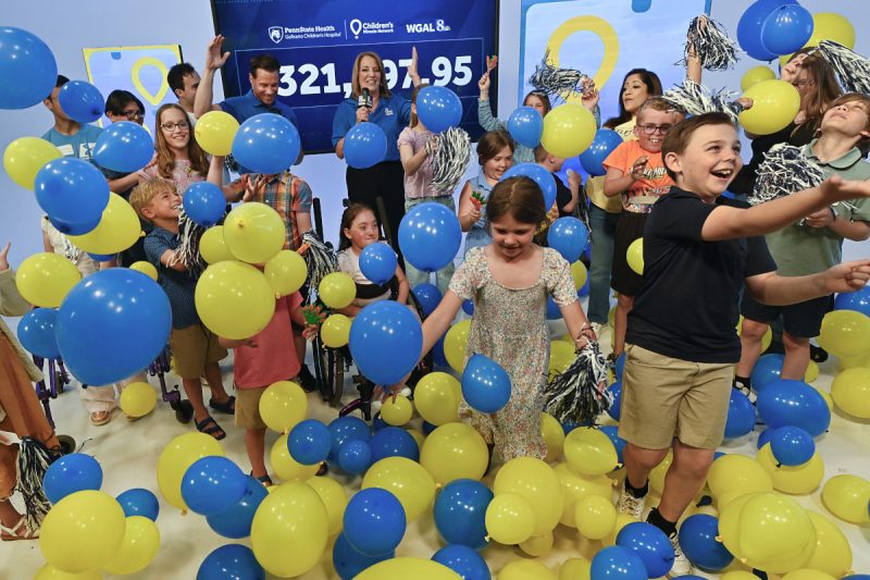 Children and event hosts celebrate during a Children’s Miracle Network fundraising reveal, tossing blue and yellow balloons into the air in front of a large display showing the total raised. The room is filled with smiling children, balloons, and pom-poms as participants cheer during the announcement.