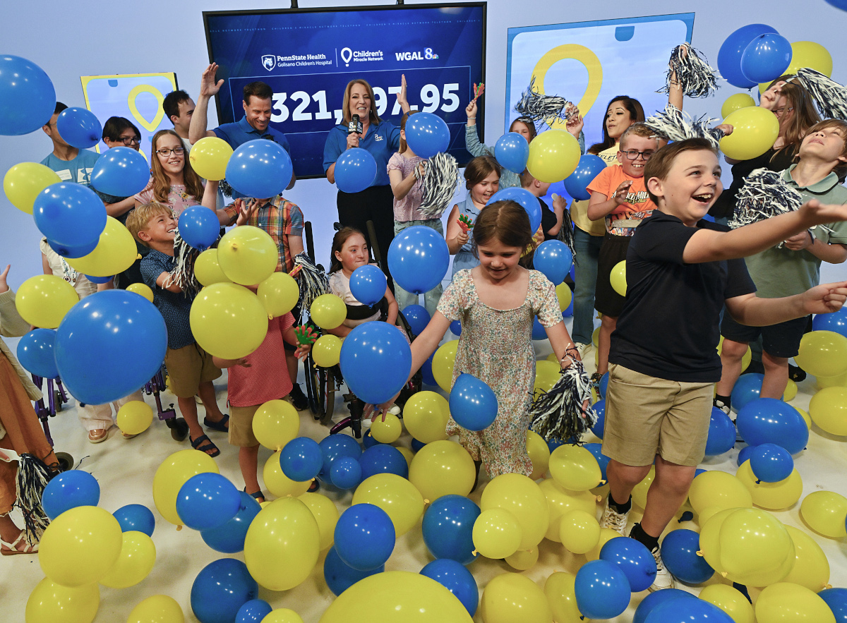 Children and event hosts celebrate during a Children’s Miracle Network fundraising reveal, tossing blue and yellow balloons into the air in front of a large display showing the total raised. The room is filled with smiling children, balloons, and pom-poms as participants cheer during the announcement.