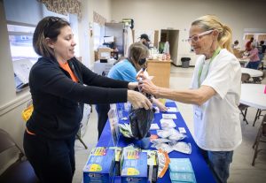 A woman holds a bag for another woman as she puts items from a table filled with brochures and other items. The woman on the left is wearnig a sweat jacket, and the woman on the right is wear a T-shirt and lanyard. Behind her is another woman who is wearing a face mask.