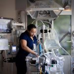 A neonatal intensive care unit (NICU) nurse adjusts equipment and cares for an infant inside an incubator. Medical monitors, IV pumps, and respiratory equipment surround the bedside, while a vital signs monitor displays the baby's heart rate and other readings.