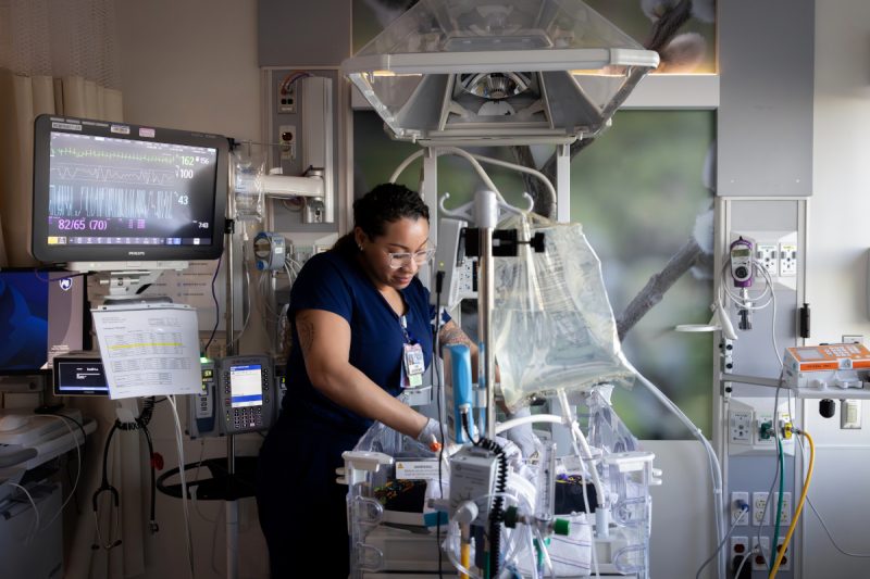A neonatal intensive care unit (NICU) nurse adjusts equipment and cares for an infant inside an incubator. Medical monitors, IV pumps, and respiratory equipment surround the bedside, while a vital signs monitor displays the baby's heart rate and other readings.