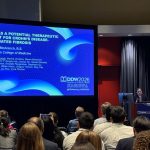 A person stands at a podium next to a large screen displaying a research presentation in front of a large conference room.