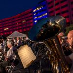 A man plays a tuba in the foreground while other musicians are in the background, in front of a crescent-shaped building lit up in red and blue lights.
