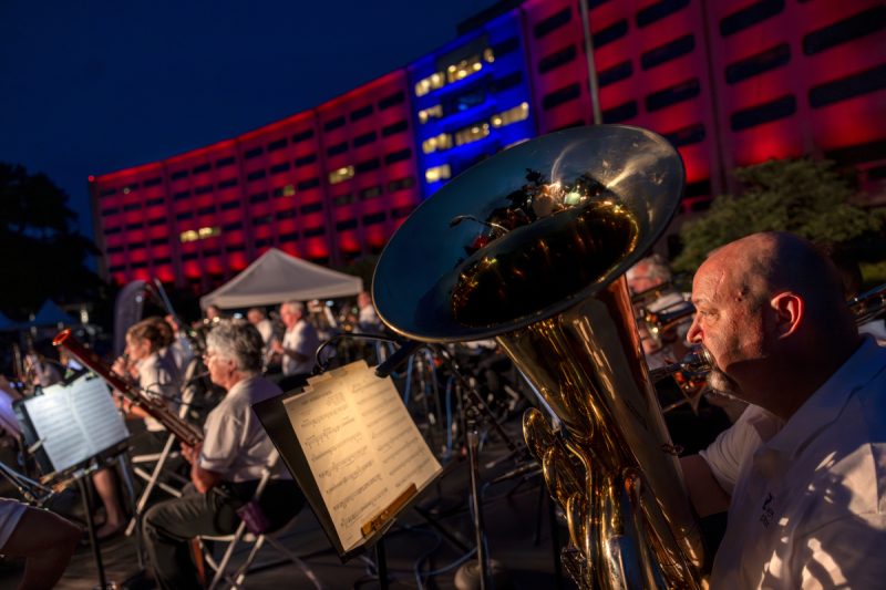 A man plays a tuba in the foreground while other musicians are in the background, in front of a crescent-shaped building lit up in red and blue lights.