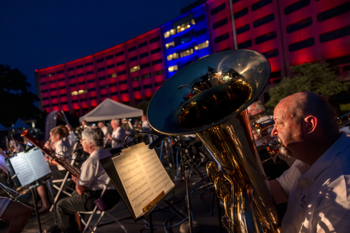A man plays a tuba in the foreground while other musicians are in the background, in front of a crescent-shaped building lit up in red and blue lights.