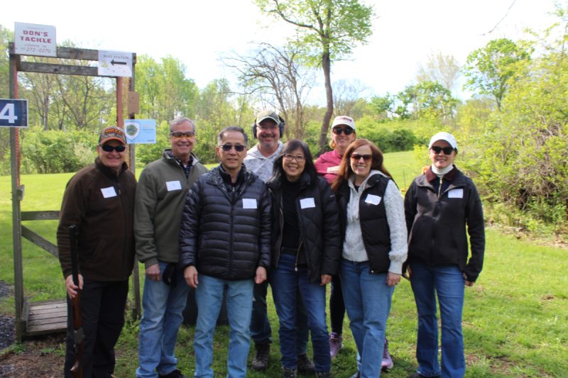 Eight people wearing casual outdoor attire pose for a photo. The man on the far left is posing with a rifle. Grass, bushes and trees are in the background.