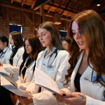 A group of students in white physician coats stand in a line holding a program.