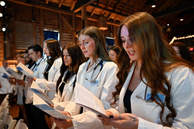 A group of students in white physician coats stand in a line holding a program.