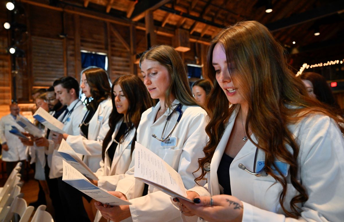A group of students in white physician coats stand in a line holding a program.