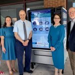 A group of people stands in front of a vending machine with a digital display screen of health items.