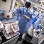 Two surgeons in blue sterile gowns and masks work in a high-tech operating room, standing beside a medical device containing a donor heart preserved in a transparent transport box while monitoring surgical equipment and screens.