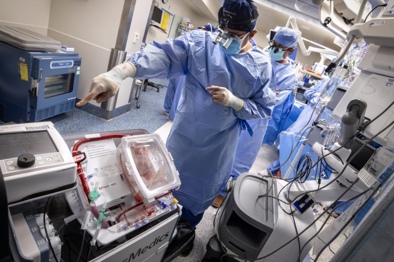 Two surgeons in blue sterile gowns and masks work in a high-tech operating room, standing beside a medical device containing a donor heart preserved in a transparent transport box while monitoring surgical equipment and screens.