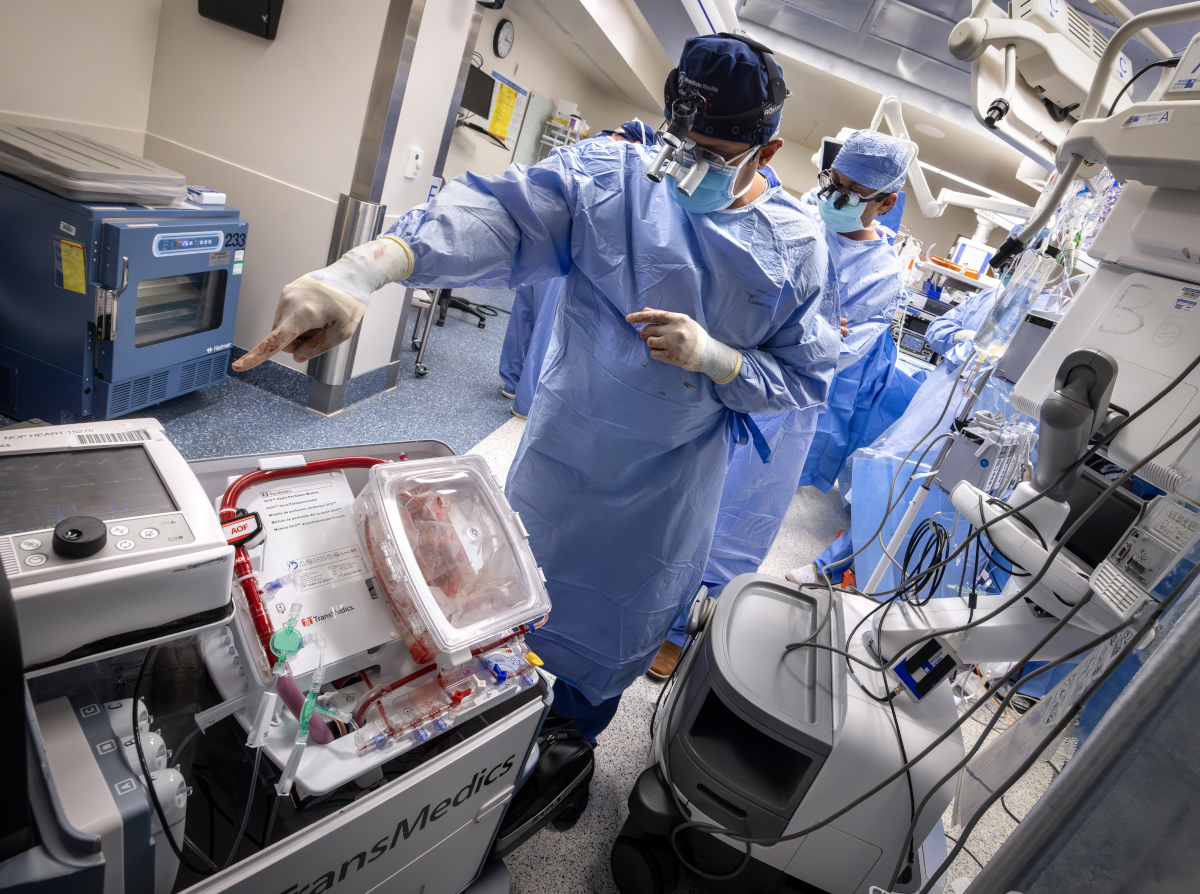 Two surgeons in blue sterile gowns and masks work in a high-tech operating room, standing beside a medical device containing a donor heart preserved in a transparent transport box while monitoring surgical equipment and screens.