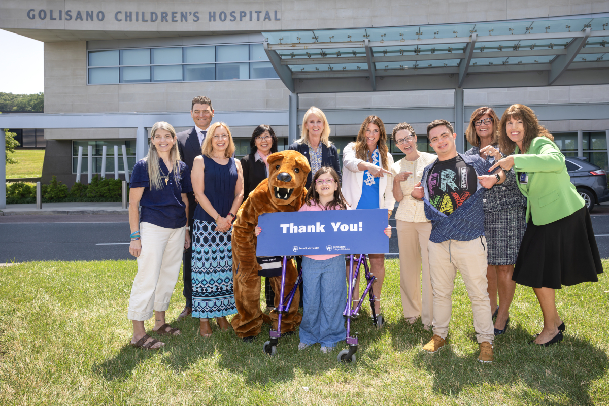 A group of people and a costumed Nittany Lion pose for a photo in front of Penn State Health Golisano Children’s Hospital, many of them smiling and pointing, “goofy pose” style. They hold a large sign that reads “Thank You!”