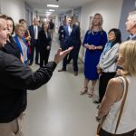 A group of about 15 people wearing professional attire stand in the hallway of a newly refurbished hospital unit. A man in the foreground has his arm extended outward as he speaks to the group.