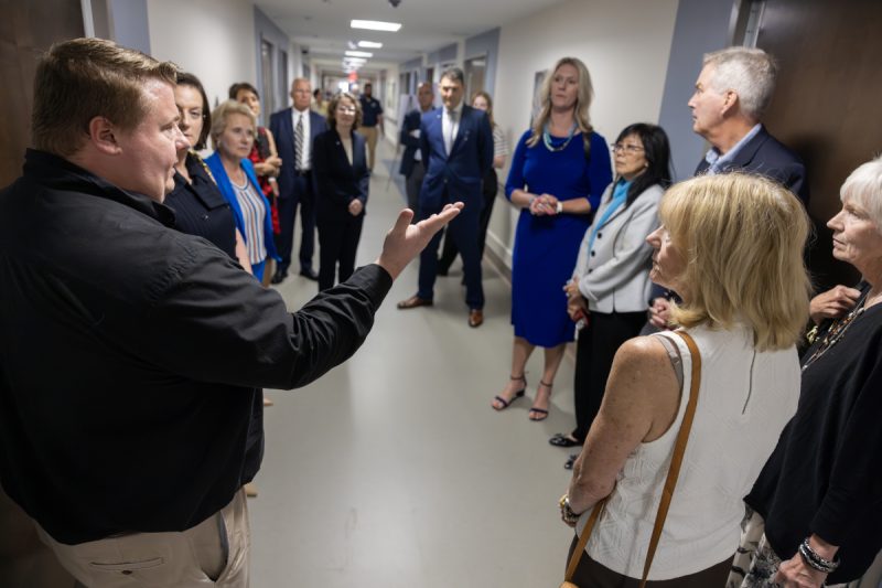 A group of about 15 people wearing professional attire stand in the hallway of a newly refurbished hospital unit. A man in the foreground has his arm extended outward as he speaks to the group.