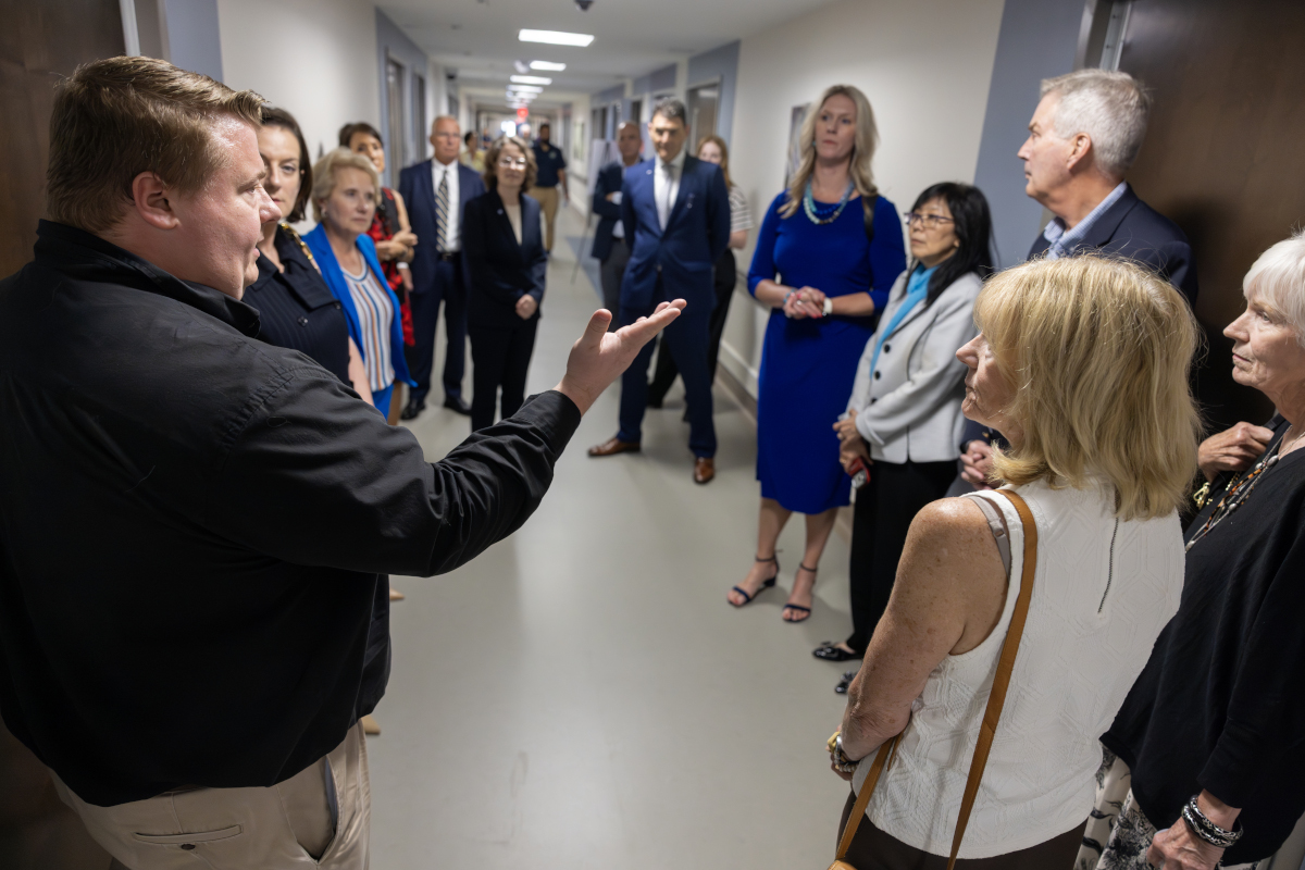 A group of about 15 people wearing professional attire stand in the hallway of a newly refurbished hospital unit. A man in the foreground has his arm extended outward as he speaks to the group.