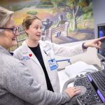 A doctor wearing a physician’s coat points to a computer screen with an ultrasound image on it. Another clinical staff member looks on. A wall in the near background has a mural of two people sitting on a bench looking out over a body of water.