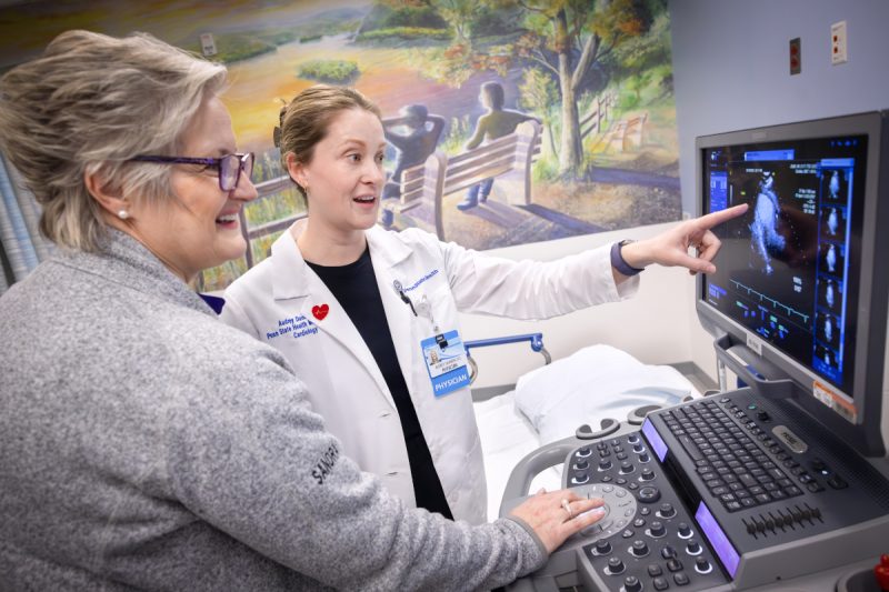 A doctor wearing a physician’s coat points to a computer screen with an ultrasound image on it. Another clinical staff member looks on. A wall in the near background has a mural of two people sitting on a bench looking out over a body of water.
