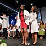 Medical students stand on a stage as faculty members help them put on their first white coats.