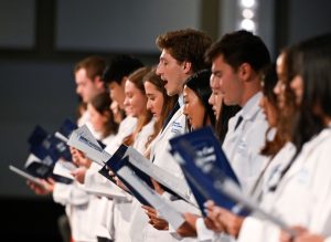 Medical students stand in a line on stage, wearing their white coats and reciting the Hippocratic Oath.