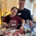 A mother and father smile in front of their infant son who is in an incubator in a hospital neonatal intensive care unit. The mother touches the baby’s hand.