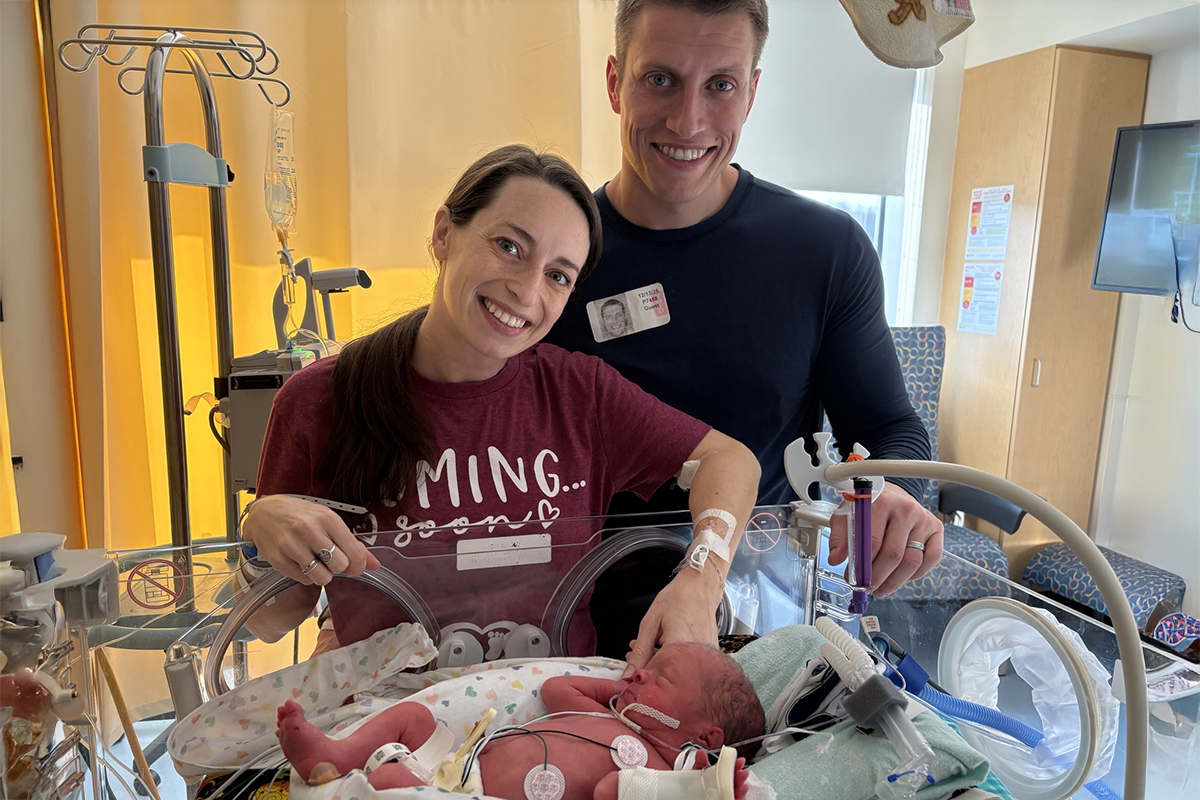 A mother and father smile in front of their infant son who is in an incubator in a hospital neonatal intensive care unit. The mother touches the baby’s hand.