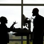 A silhouette of two employees standing and talking in front of a window at Penn State Health St. Joseph Medical Center. A computer on a cart is between the two of them.