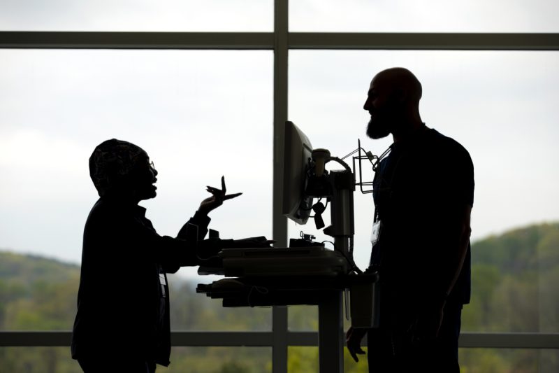A silhouette of two employees standing and talking in front of a window at Penn State Health St. Joseph Medical Center. A computer on a cart is between the two of them.