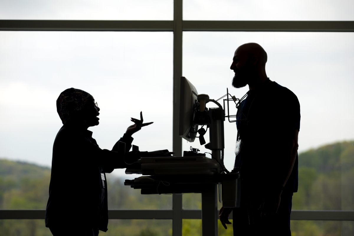 A silhouette of two employees standing and talking in front of a window at Penn State Health St. Joseph Medical Center. A computer on a cart is between the two of them.