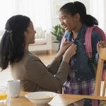 Mother buttons daughter’s jacket as she gets ready for school. Daughter is wearing a backpack. A table with breakfast dishes is in front of them.