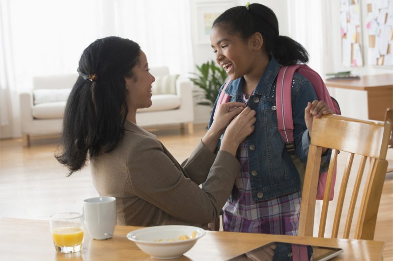 Mother buttons daughter’s jacket as she gets ready for school. Daughter is wearing a backpack. A table with breakfast dishes is in front of them.