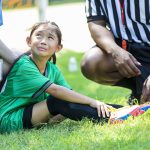 Young female soccer player holds her ankle and grimaces in pain while crying. She is sitting on the playing field with her coach and referee gathered around her.