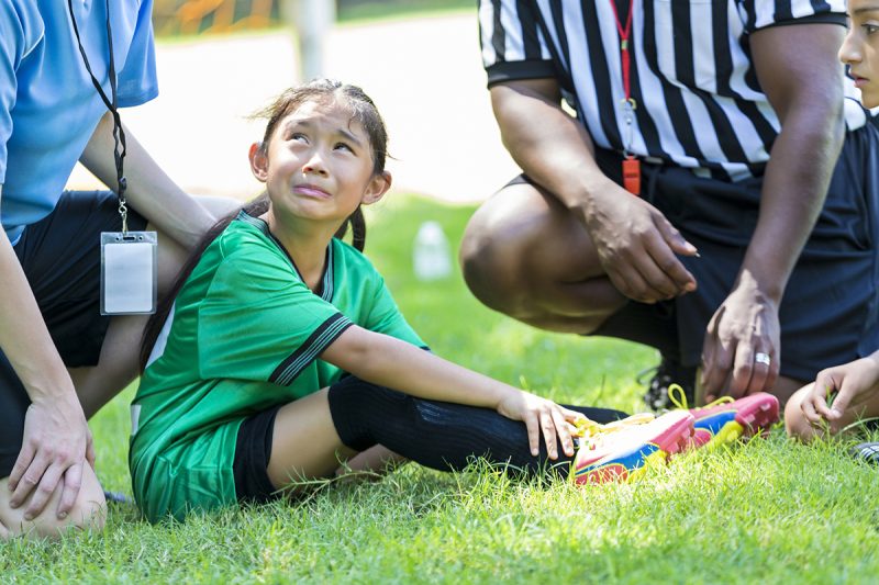 Young female soccer player holds her ankle and grimaces in pain while crying. She is sitting on the playing field with her coach and referee gathered around her.