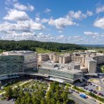 Aerial view of the Penn State Health Milton S. Hershey Medical Center campus with multiple modern buildings, parking areas, landscaped grounds, and a helipad, surrounded by wooded hills and farmland under a partly cloudy blue sky.